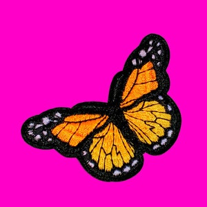 May include: Embroidered butterfly patch with orange and yellow wings, outlined in black. The patch has white accents on the wing edges and is set against a bright pink background. This decorative patch is suitable for clothing or accessories.