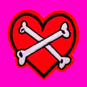 May include: Red heart-shaped patch with a white embroidered crossbones design. The heart has a black outline.