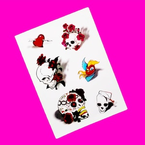 May include: A sheet of temporary tattoos featuring various skull designs with floral accents. The designs include a heart with an arrow, a skull with a crown of flowers, a skull with a floral design, a skull with a floral design and a bird, and a skull with a floral design and a heart.