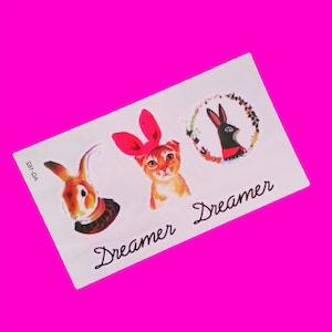 May include: Three temporary tattoos featuring a cat with pink bunny ears, a rabbit with a white collar, and a black rabbit with a red bow in a wreath of flowers. The tattoos are on a white background with the word "Dreamer" written twice.