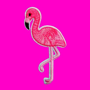 Könnte beinhalten: Gestickter pinkfarbener Flamingo-Aufnäher mit weißem Rand, der auf einem Bein steht. Der Flamingo hat einen schwarzen Schnabel und ein Auge, vor einem leuchtend pinkfarbenen Hintergrund. Ideal zum Aufnähen auf Kleidung oder Accessoires.