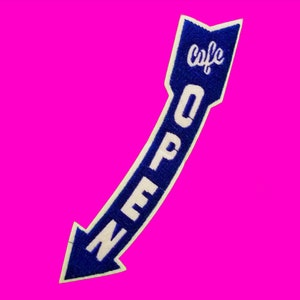 Puede incluir: Un parche bordado azul y blanco en forma de flecha, con la palabra "OPEN" y "cafe" en letras blancas. El parche tiene un borde blanco y está sobre un fondo rosa brillante.