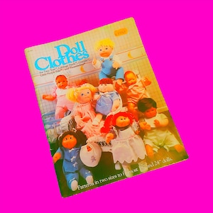May include: A book titled "Doll Clothes" with a colorful cover featuring a group of dolls wearing various outfits. The book includes patterns for making clothes for dolls that are 18 inches and 24 inches tall.