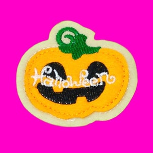 Puede incluir: Una calabaza de fieltro amarilla con un tallo verde y un "Halloween" bordado en blanco en la parte delantera. La calabaza tiene una cara bordada negra con dos ojos y una amplia sonrisa.