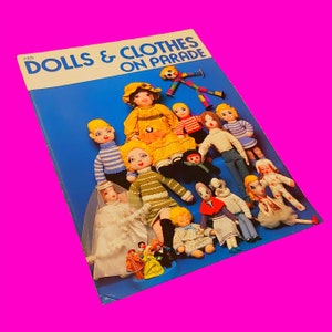 May include: A blue book titled "DOLLS & CLOTHES ON PARADE" featuring a collection of handmade dolls. The dolls are dressed in diverse outfits, including a bride, a yellow dress, and striped sweaters. The background is a bright pink.