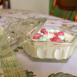 May include: A clear glass candle holder with a lid, filled with a white candle and decorated with pink and green candy-shaped wax pieces. The candle holder is hexagonal and sits on a floral patterned surface.