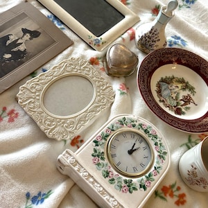 May include: An assortment of vintage decorative items, including picture frames, a heart-shaped trinket box, a small clock, a decorative plate, and a perfume bottle. The items are in various colors, including cream, gold, and burgundy, and are arranged on a floral patterned blanket.