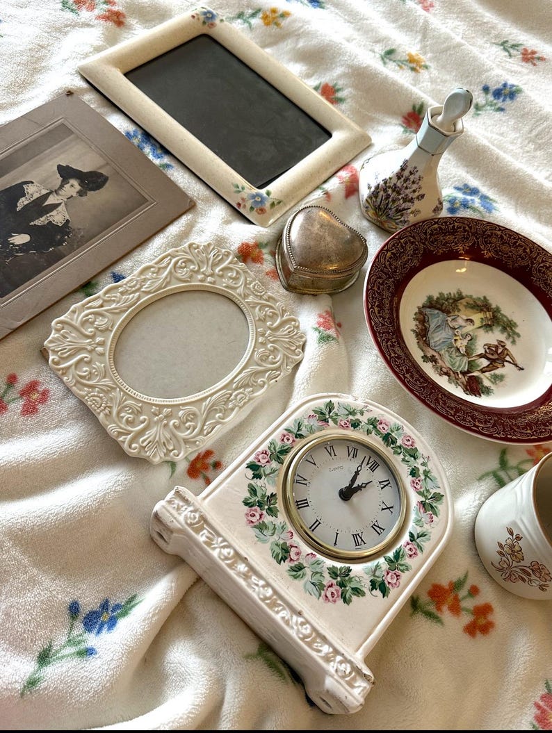 May include: An assortment of vintage decorative items, including picture frames, a heart-shaped trinket box, a small clock, a decorative plate, and a perfume bottle. The items are in various colors, including cream, gold, and burgundy, and are arranged on a floral patterned blanket.