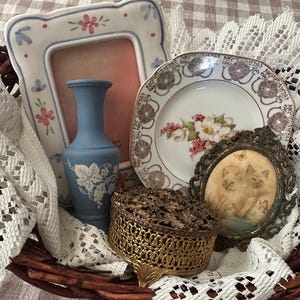 May include: A wicker basket holds a collection of vintage items. A blue vase with a grape design, a floral-decorated plate, a small framed picture of cats, and a decorative gold box are visible. White lace and a floral-framed picture complete the arrangement.