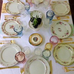 May include: A white tablecloth with a variety of gold-rimmed plates, gold silverware, and colorful glassware. The plates have floral patterns and the glassware is clear, blue, and pink.