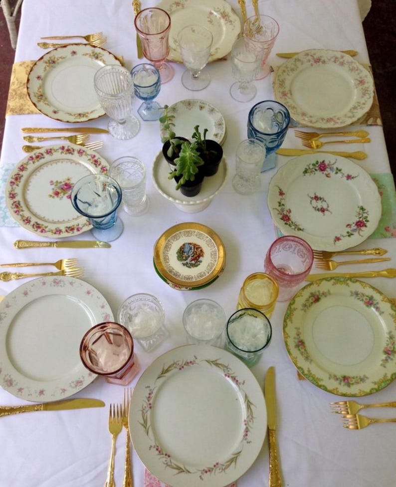 May include: A white tablecloth with a variety of gold-rimmed plates, gold silverware, and colorful glassware. The plates have floral patterns and the glassware is clear, blue, and pink.