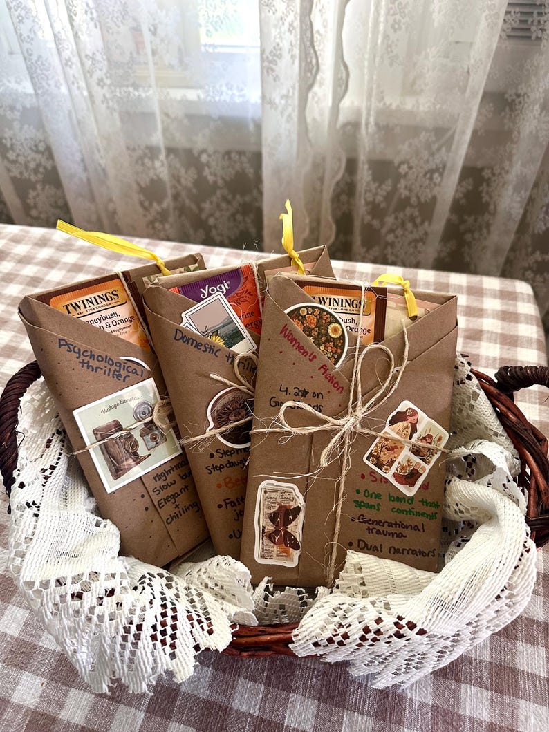 May include: A wicker basket holds several brown paper-wrapped packages, each tied with a yellow ribbon. The packages contain tea and are labeled with handwritten text and decorative stickers. The labels include the words "Psychological thriller", "Domestic", and "Women's Fiction".