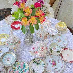 May include: A collection of vintage teacups, saucers, and teapots with floral patterns, arranged on a white crocheted tablecloth with a bouquet of pink, white, and orange flowers in a glass vase.