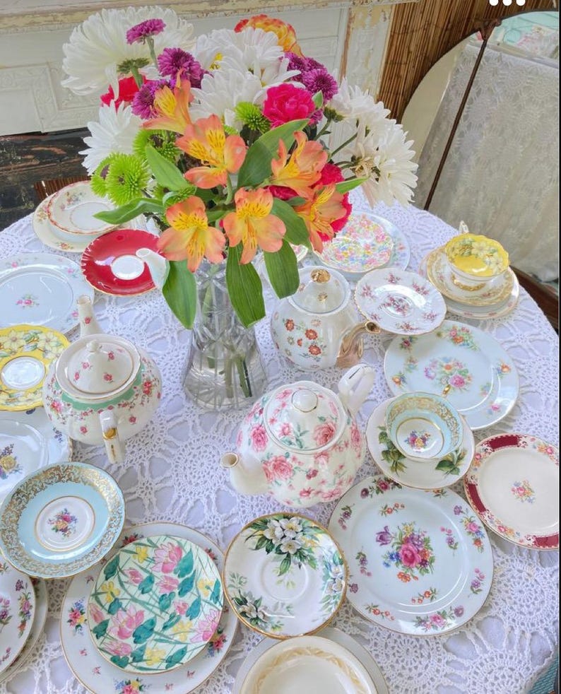 May include: A collection of vintage teacups, saucers, and teapots with floral patterns, arranged on a white crocheted tablecloth with a bouquet of pink, white, and orange flowers in a glass vase.