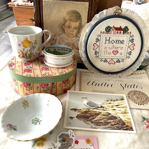May include: A collection of vintage items, including a floral teacup, a small round trinket box, a cross-stitch hoop with the text "Home is where the is", a floral patterned box, a postcard with a seagull, and a small card that says "Thank You!!".