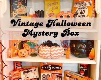 Puede incluir: Una estantería blanca llena de artículos vintage de Halloween. La estantería muestra cajas de caramelos, decoraciones de calabazas y una máscara de gato negro. El texto "Vintage Halloween Mystery Box" está en la imagen.