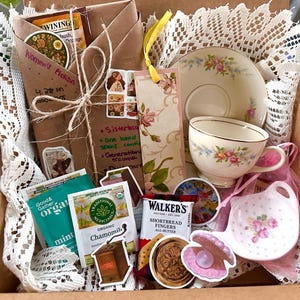 May include: A gift box filled with a teacup and saucer set, tea bags, a bookmark, and a small dish. The teacup and saucer have a floral design. The box also contains a wrapped book and stickers.