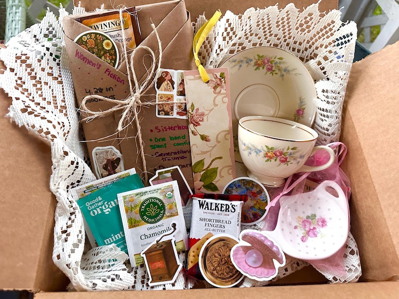 May include: A gift box filled with a teacup and saucer set, tea bags, a bookmark, and a small dish. The teacup and saucer have a floral design. The box also contains a wrapped book and stickers.