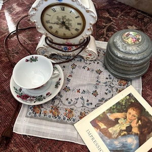 May include: A collection of vintage items including a white porcelain clock with floral accents, a teacup and saucer set, a small metal trinket box, a floral handkerchief, and a book titled "Impressionism". The clock has a round face with Roman numerals.