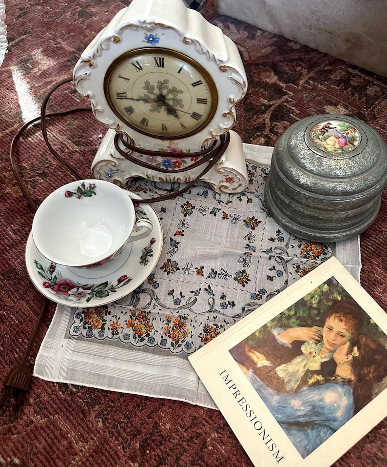 May include: A collection of vintage items including a white porcelain clock with floral accents, a teacup and saucer set, a small metal trinket box, a floral handkerchief, and a book titled "Impressionism". The clock has a round face with Roman numerals.