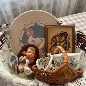 May include: A collection of vintage decorative items, including a plate with a goose design, a framed picture of children, and a small basket with a teacup. The items are arranged on a lace cloth.