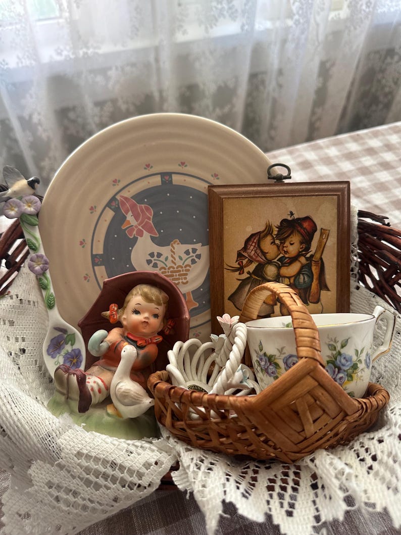 May include: A collection of vintage decorative items, including a plate with a goose design, a framed picture of children, and a small basket with a teacup. The items are arranged on a lace cloth.