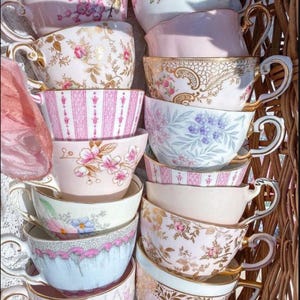 May include: A stack of vintage teacups with floral and patterned designs in shades of pink, blue, and white. The cups are arranged in a wicker basket.