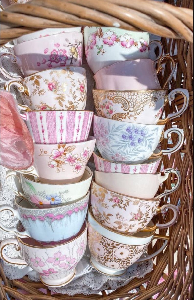 May include: A stack of vintage teacups with floral and patterned designs in shades of pink, blue, and white. The cups are arranged in a wicker basket.