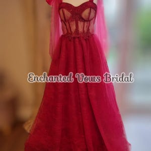 May include: A red tulle wedding dress with a lace bodice and a sweetheart neckline. The dress has a fitted bodice and a flowing skirt. The dress is on a mannequin and has a bow on each shoulder strap. The text "Enchanted Vows Bridal" is visible on the dress.