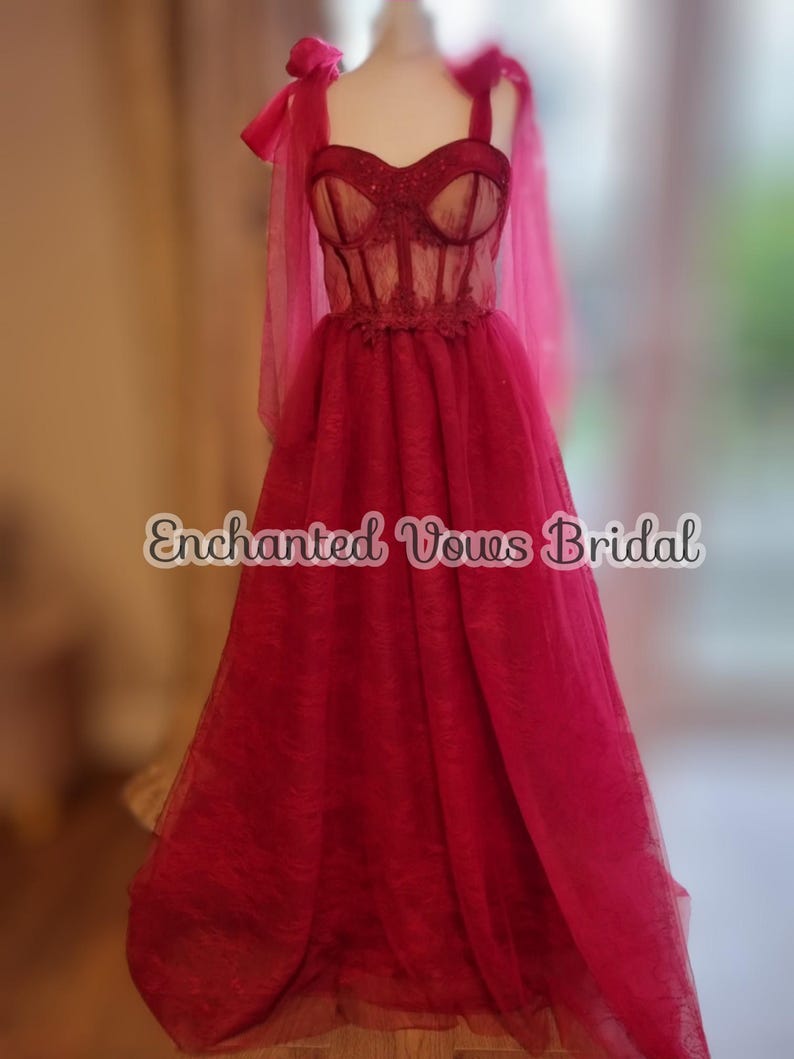 May include: A red tulle wedding dress with a lace bodice and a sweetheart neckline. The dress has a fitted bodice and a flowing skirt. The dress is on a mannequin and has a bow on each shoulder strap. The text "Enchanted Vows Bridal" is visible on the dress.