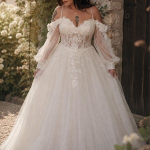 May include: An ivory floor-length gown featuring a sweetheart neckline, sheer corset-style bodice with floral lace appliqués, and long, loose-fitting sheer sleeves. The full skirt is made of layered tulle with delicate floral embroidery cascading down.