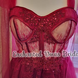 May include: A red tulle wedding dress with a lace bodice and a sweetheart neckline. The bodice features a floral design with sequins. The dress has a flowing skirt and a train. The text "Enchanted Vows Bridal" is visible on the dress.