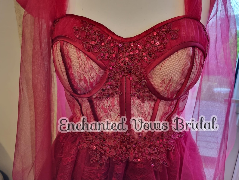 May include: A red tulle wedding dress with a lace bodice and a sweetheart neckline. The bodice features a floral design with sequins. The dress has a flowing skirt and a train. The text "Enchanted Vows Bridal" is visible on the dress.