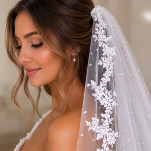 May include: A white bridal veil with delicate floral lace detailing and scattered pearl accents. The veil is attached to the hair and drapes gracefully. The bride is wearing a pearl earring.