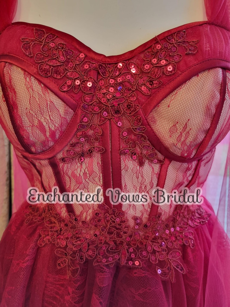 May include: A red lace and tulle wedding dress with a sweetheart neckline and a corset bodice. The bodice is decorated with red sequins and floral embroidery. The skirt is full and flowing. The dress is labeled "Enchanted Vows Bridal".