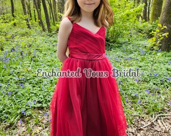 May include: A young girl wearing a red tulle dress with a satin sash and lace trim. The dress has a V-neckline and spaghetti straps. The girl is standing in a woodland setting with blue flowers in the background. The text "Enchanted Vows Bridal" is visible in the image.