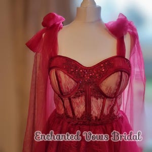 May include: A red lace and tulle wedding dress with a sweetheart neckline and a corset bodice. The dress has a bow on the shoulder strap and the text "Enchanted Vows Bridal" is printed on the skirt.