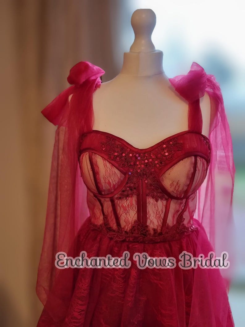 May include: A red lace and tulle wedding dress with a sweetheart neckline and a corset bodice. The dress has a bow on the shoulder strap and the text "Enchanted Vows Bridal" is printed on the skirt.
