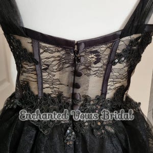 May include: Black tulle wedding dress with lace and sequin embellishments. The dress has a fitted bodice with a corset-style back and a full skirt. The back of the dress features a row of buttons.