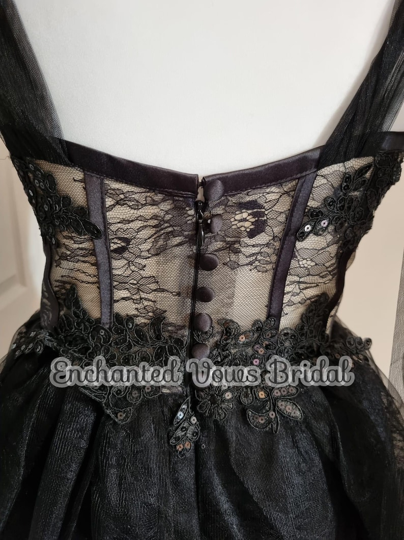 May include: Black tulle wedding dress with lace and sequin embellishments. The dress has a fitted bodice with a corset-style back and a full skirt. The back of the dress features a row of buttons.