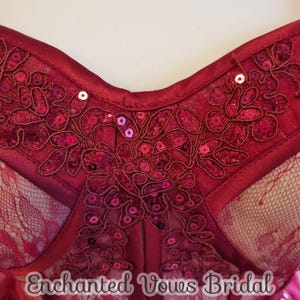 May include: Close-up of a burgundy corset with lace and sequins. The corset has a sweetheart neckline and is adorned with floral embroidery. The text "Enchanted Vows Bridal" is visible at the bottom of the image.