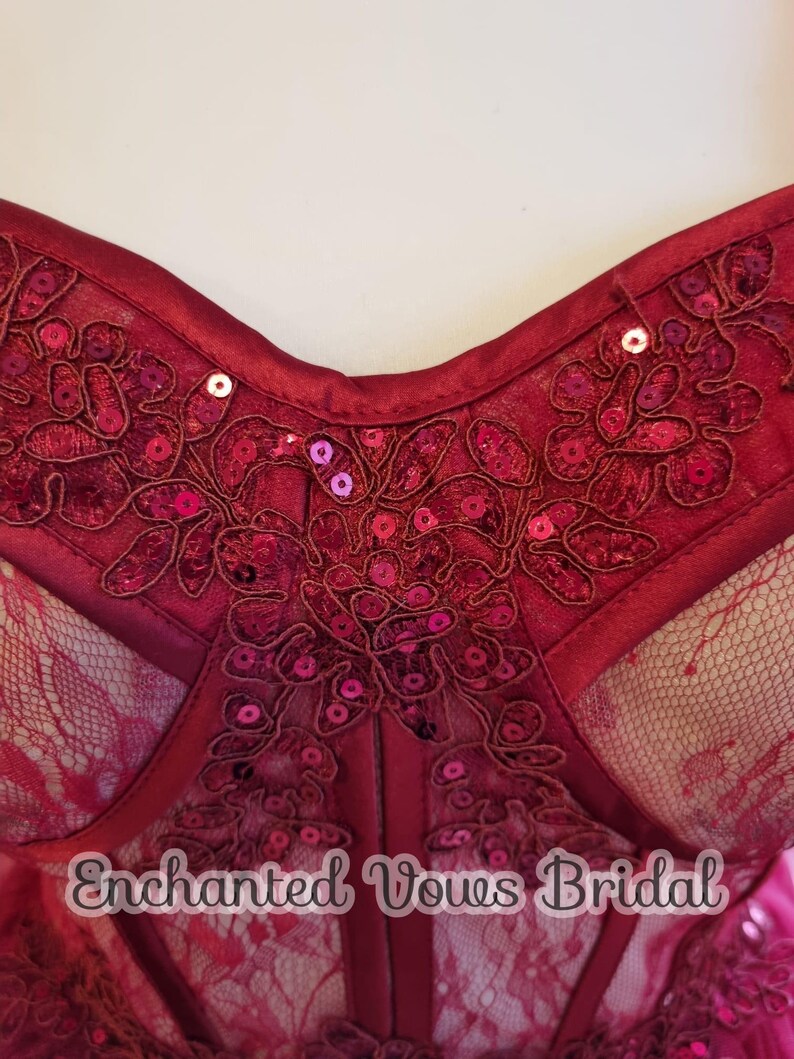 May include: Close-up of a burgundy corset with lace and sequins. The corset has a sweetheart neckline and is adorned with floral embroidery. The text "Enchanted Vows Bridal" is visible at the bottom of the image.