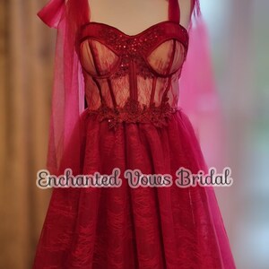 May include: A red tulle wedding dress with a lace bodice and a bow on the shoulder strap. The dress is on a mannequin and the text "Enchanted Vows Bridal" is visible at the bottom of the image.