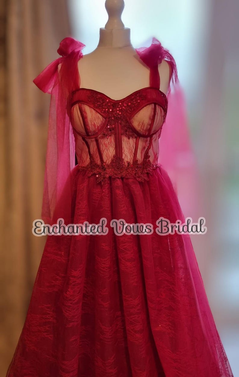 May include: A red tulle wedding dress with a lace bodice and a bow on the shoulder strap. The dress is on a mannequin and the text "Enchanted Vows Bridal" is visible at the bottom of the image.