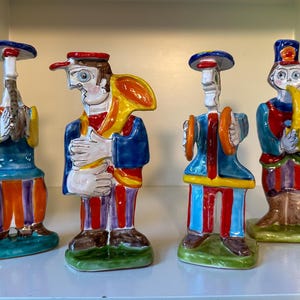 May include: Four colorful ceramic figurines of musicians. Each figurine is wearing a different hat and playing a different instrument. The instruments include a trumpet, a saxophone, a drum, and a tambourine.