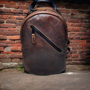 Puede incluir: Una mochila de cuero marrón con una cremallera negra y una correa negra. La mochila está sentada sobre una superficie de concreto frente a una pared de ladrillo.