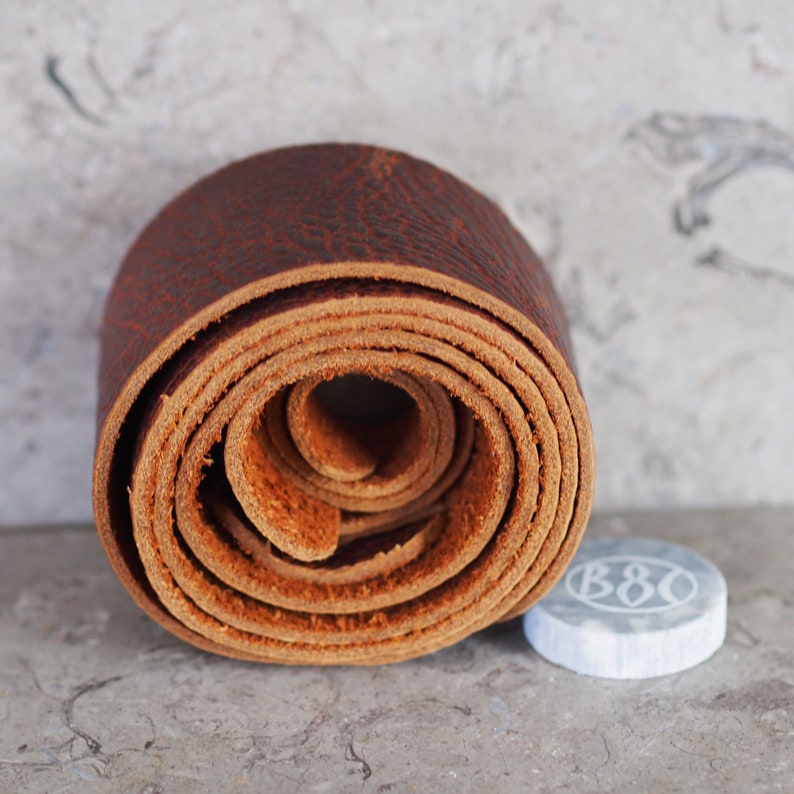 May include: A close-up of a rolled up brown leather strap. The strap is rolled into a coil shape and has a natural, rustic look.