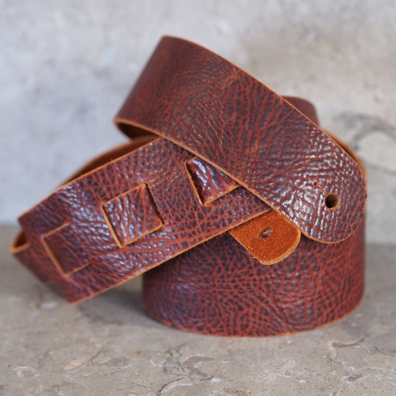 May include: A brown leather guitar strap with a textured surface and a unique design featuring cut-out squares. The strap is folded in half, showing the inside of the strap.