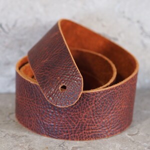 May include: A brown leather guitar strap with a textured surface. The strap is folded in half and the ends are overlapping.