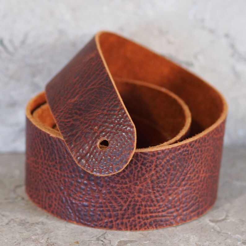 May include: A brown leather guitar strap with a textured surface. The strap is folded in half and the ends are overlapping.